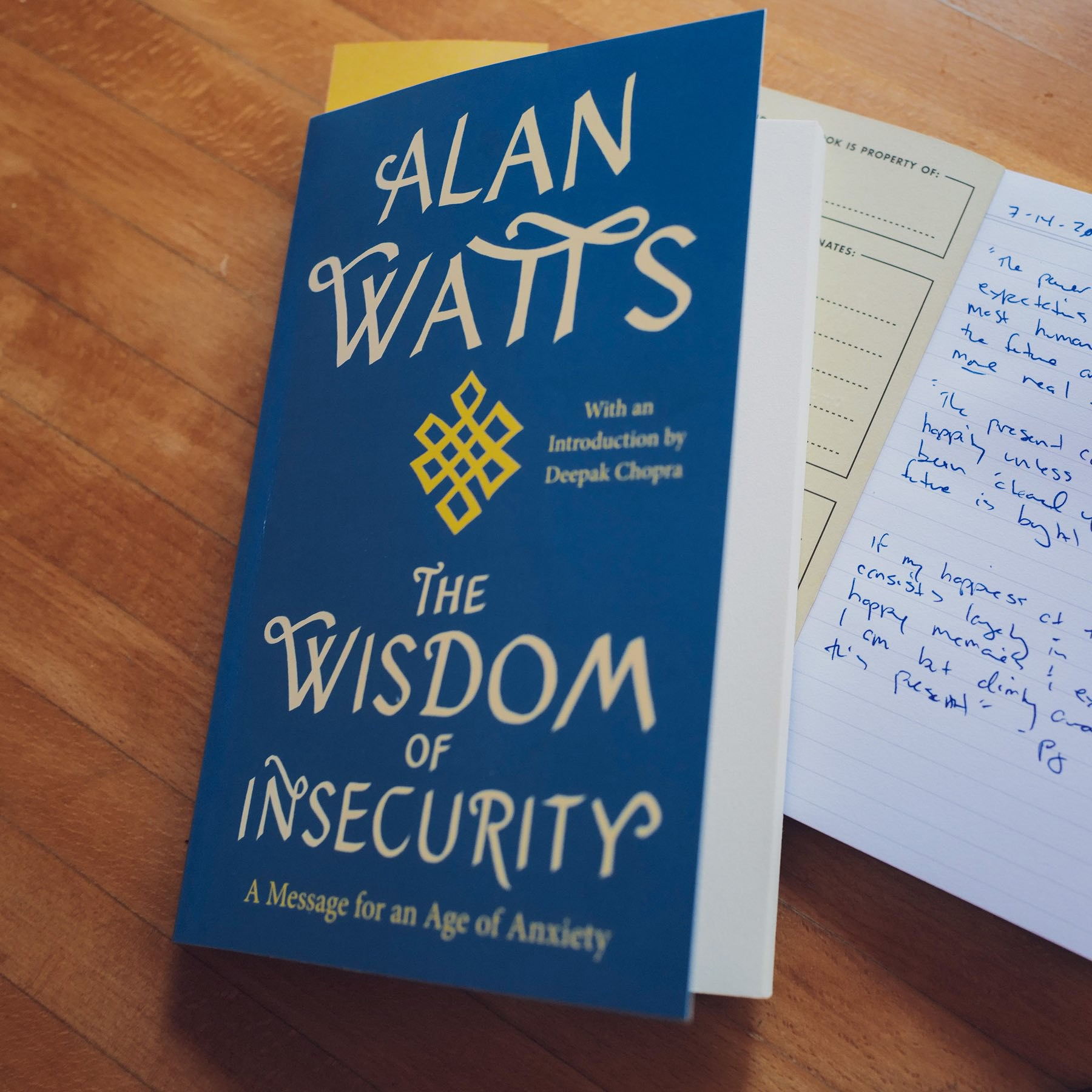 Alan Watts' book "The Wisdom of Insecurity" on a wooden table with an open notebook next to it.