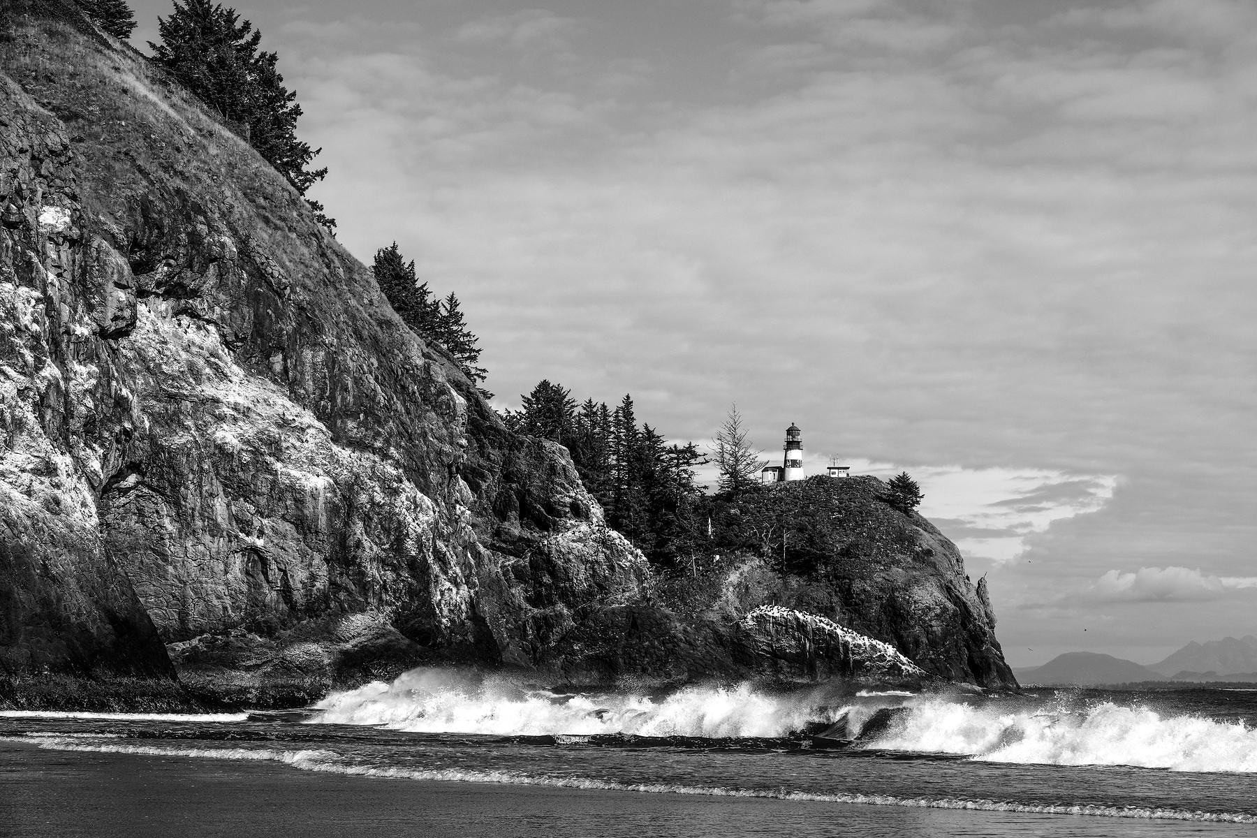 Monochrome. Waves crash against the cliffs beneath a lighthouse.