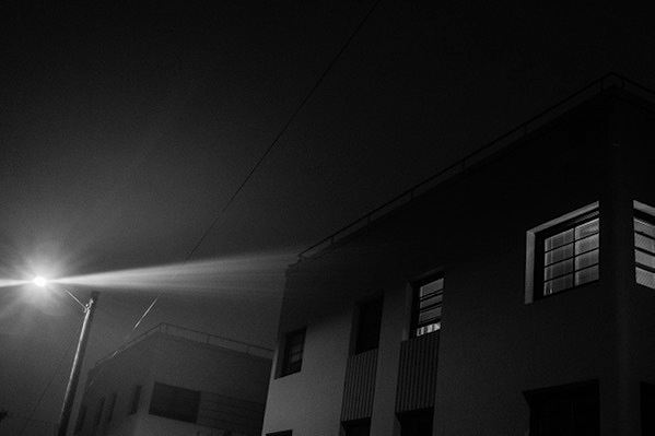 Monochrome image of lights shining through an apartment window. The shot is at a canted angle.