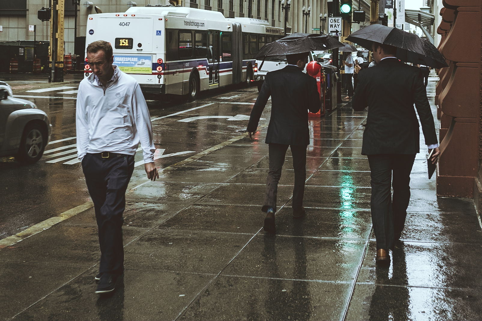 A man walks down a windy Chicago sidewalk squinting into the rain. People around him carry umbrellas.