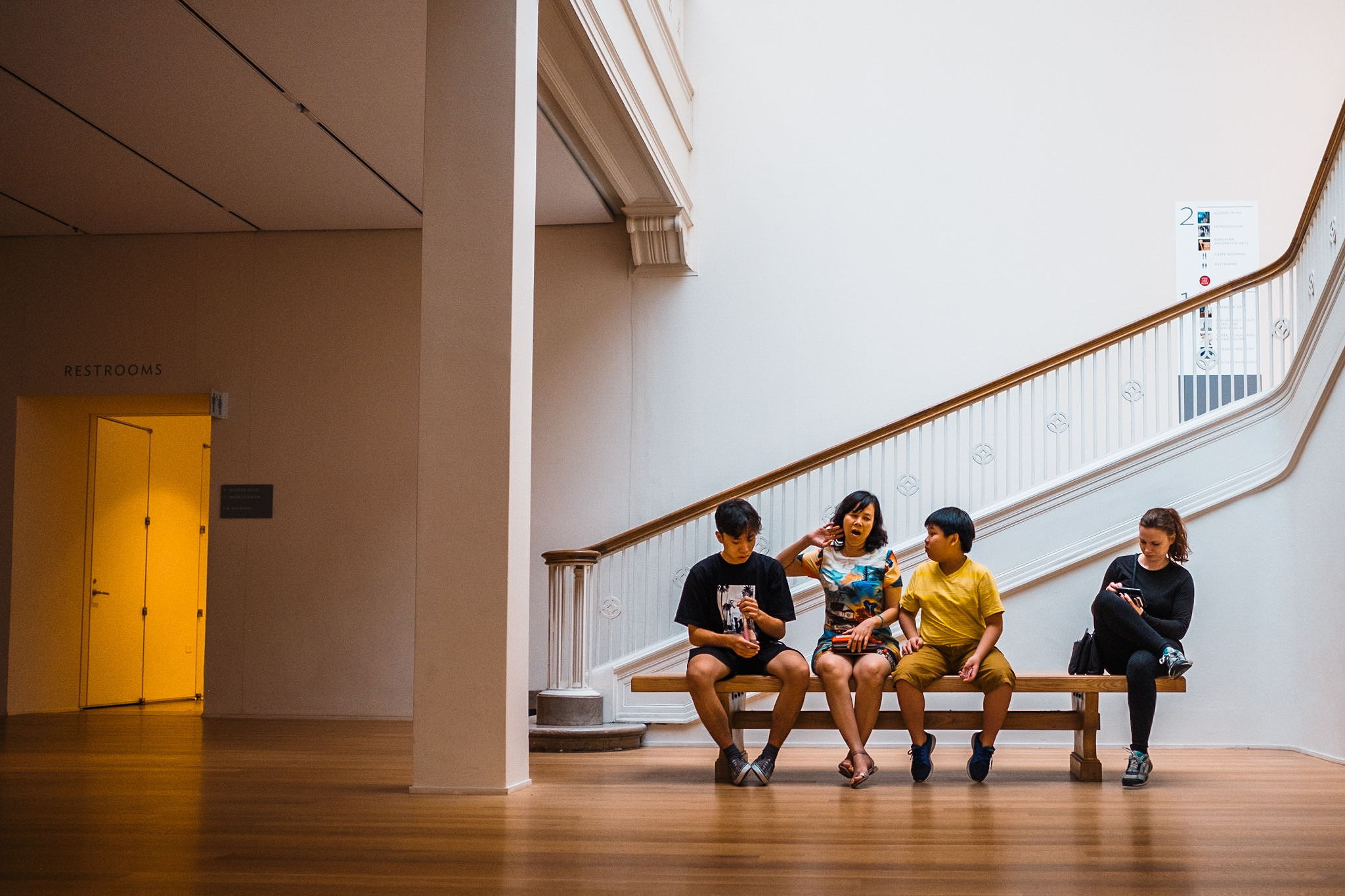 A family sits on a bench in a museum. They appear to be in animated conversation, but one of them is looking away as if to ignore what she's hearing.