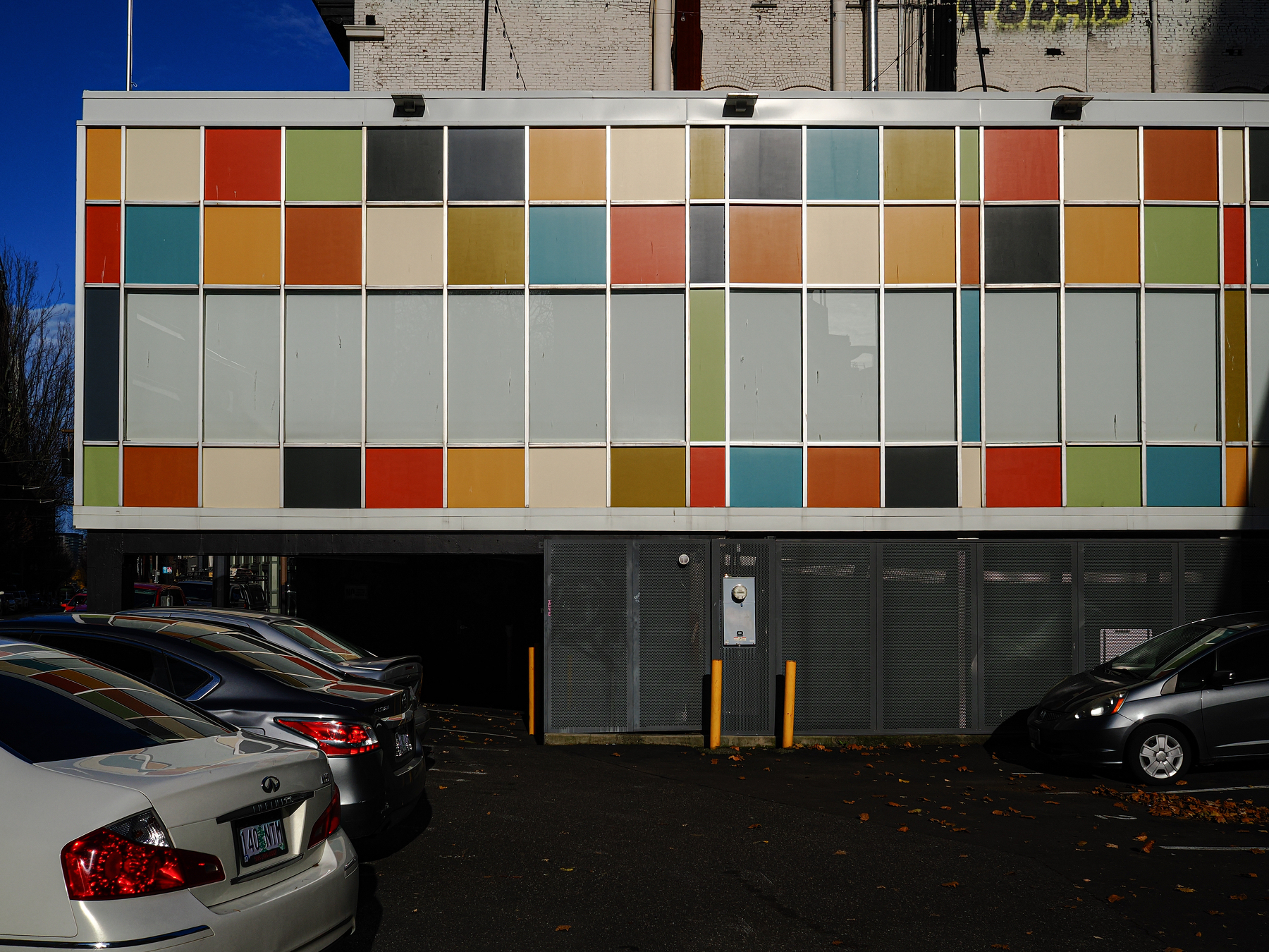 A building exterior features a grid of colorful rectangular panels above a row of parked cars in a shadowed area.