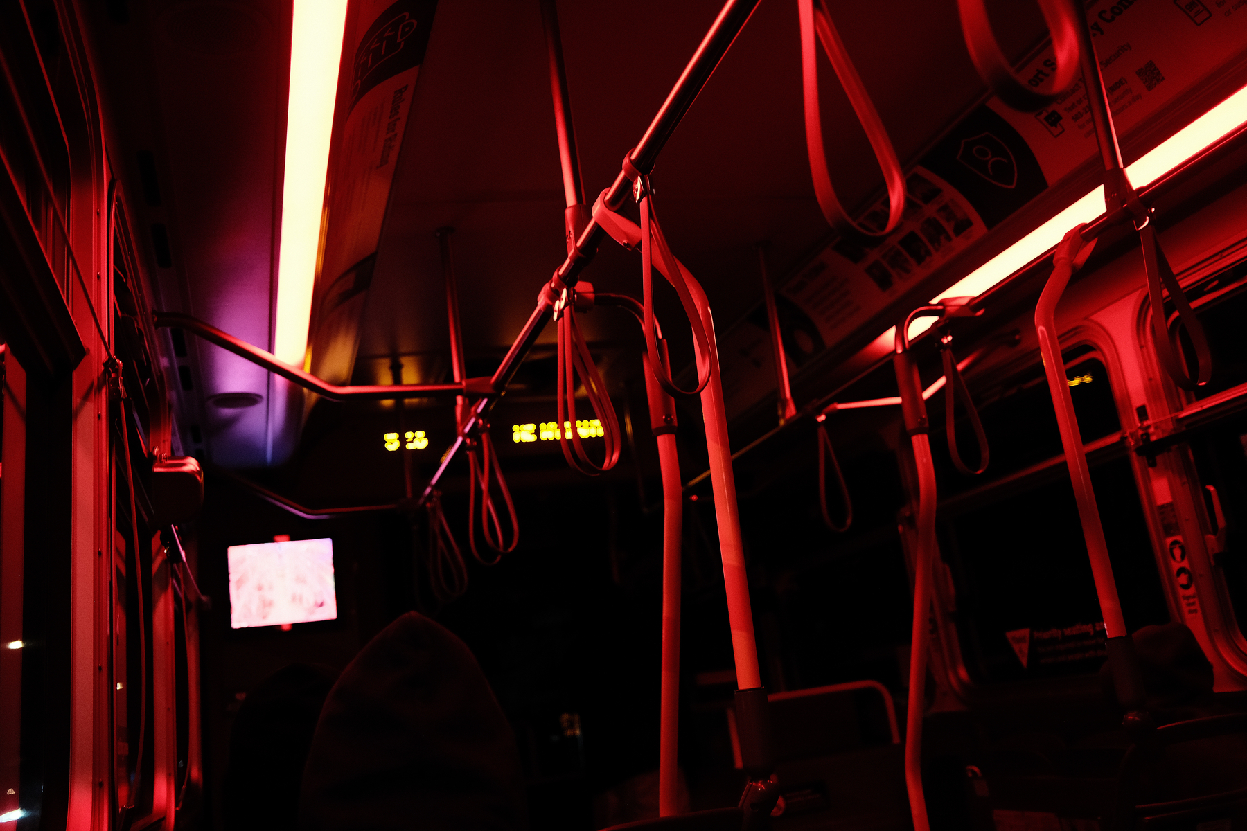 A dimly lit bus interior is illuminated by red lights with empty seats and handrails.