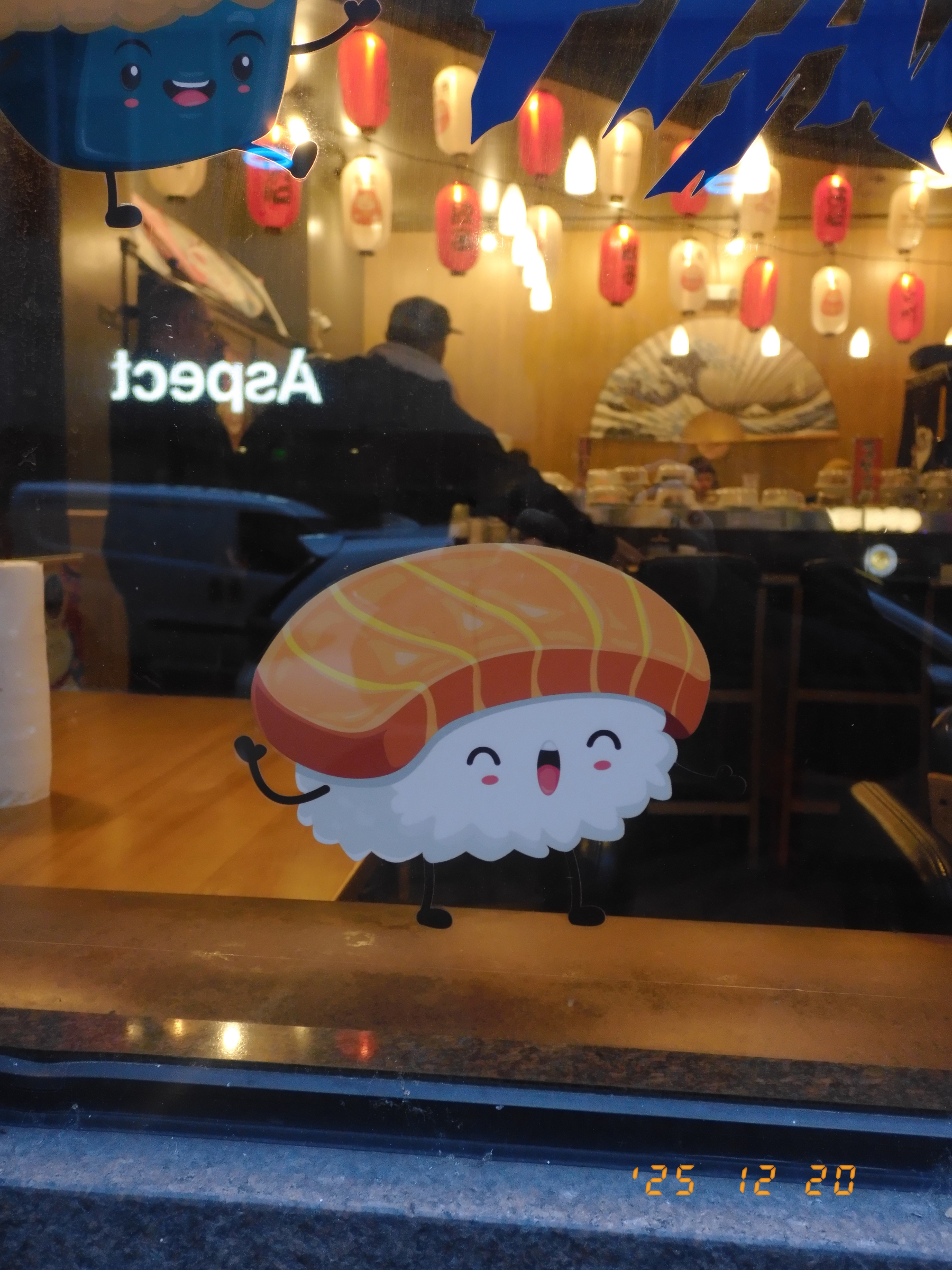 A cute cartoon sushi character is displayed on a restaurant window with people dining and lanterns visible inside.