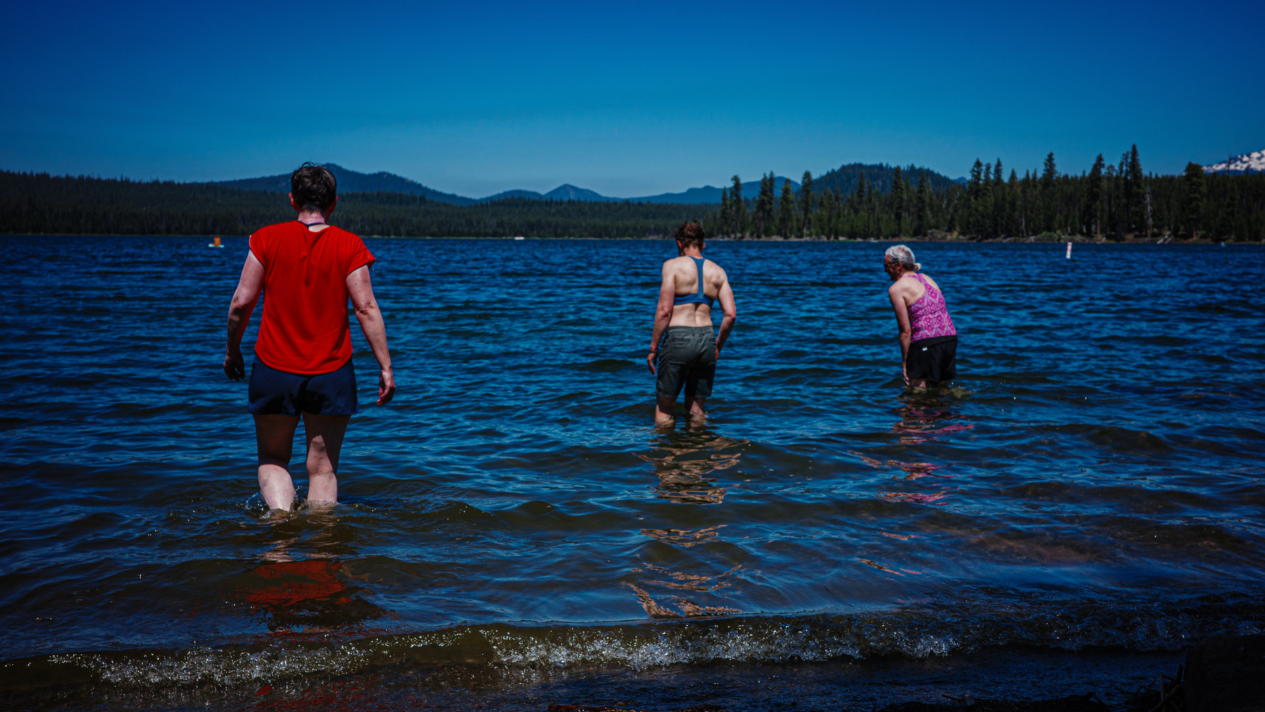 Three people are wading into a lake with a forested shoreline and mountains in the background.