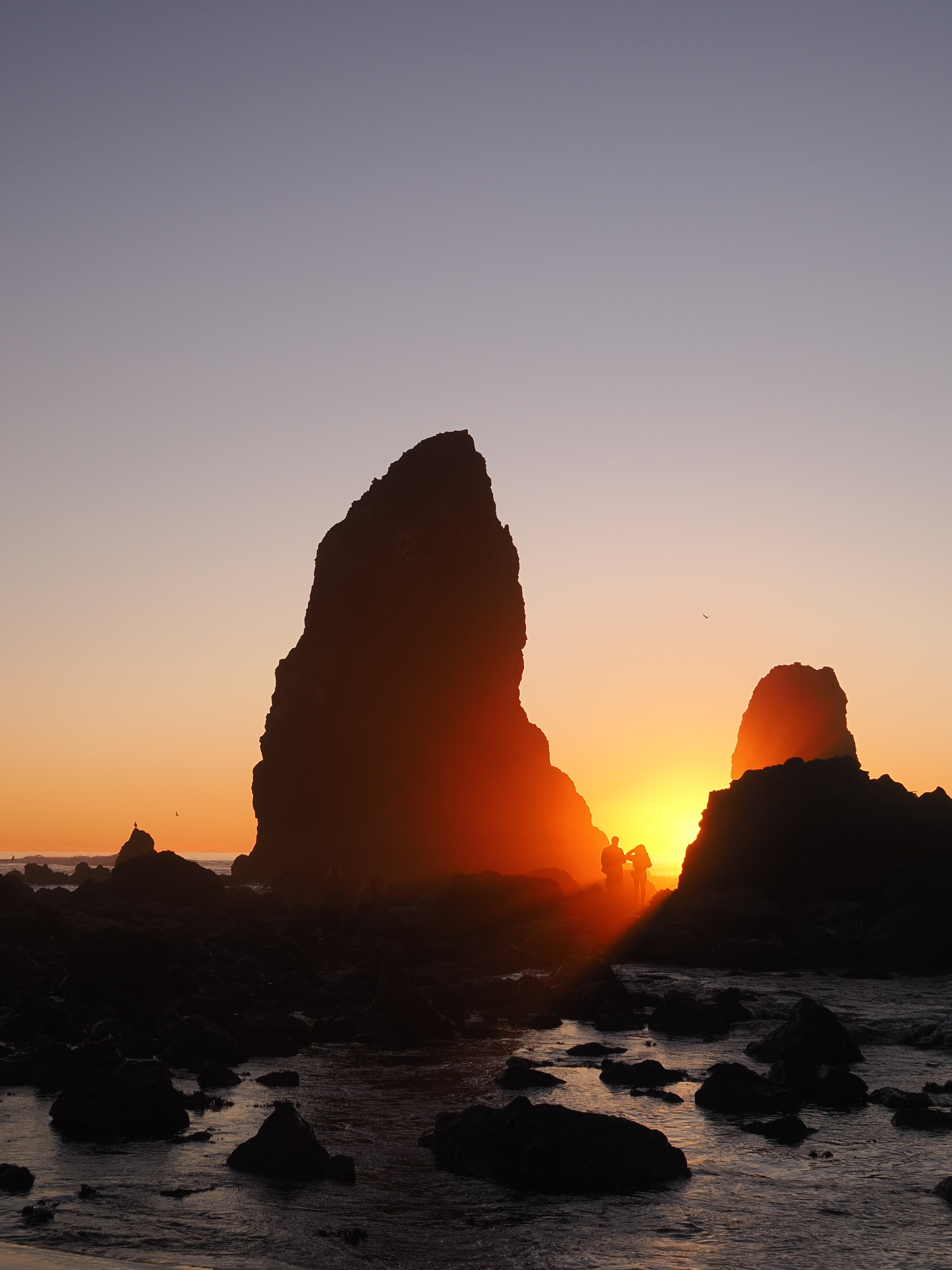 A dramatic sunset illuminates the ocean and large silhouetted rock formations.