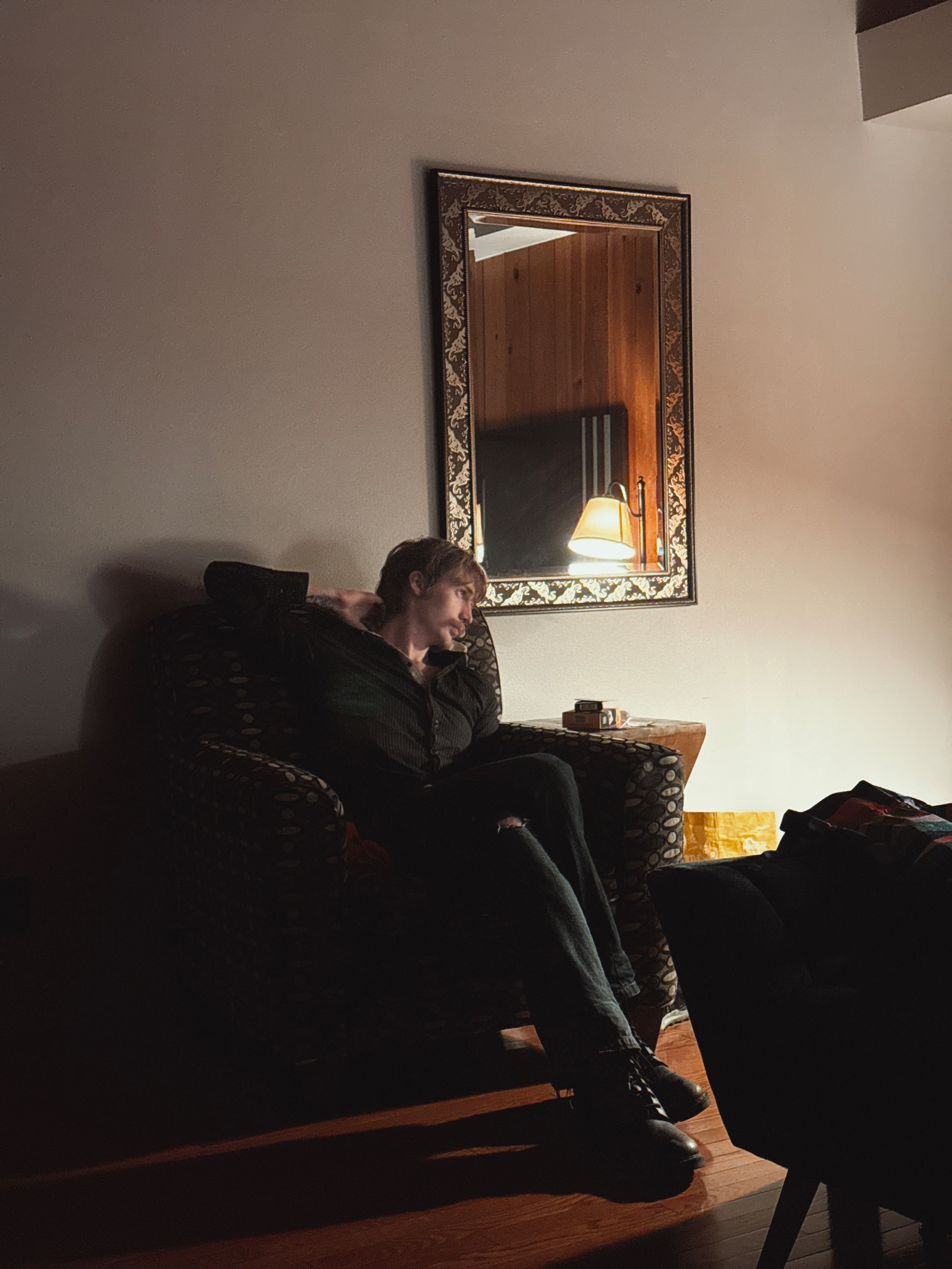 A person is sitting in a dimly lit room with a framed mirror reflecting a lamplit interior.
