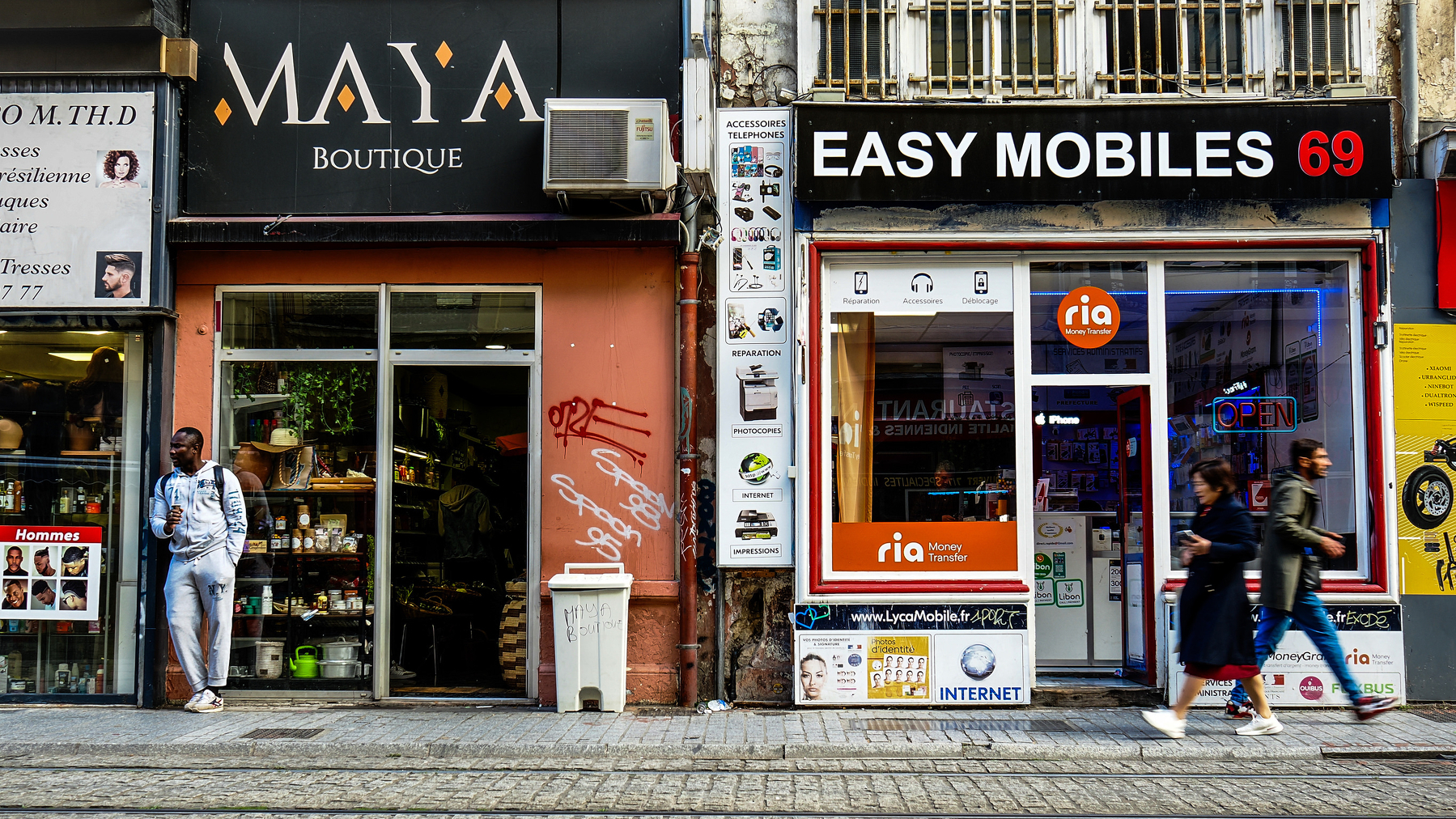 Two people walk past storefronts labeled Maya Boutique and Easy Mobiles 69 on a busy street.