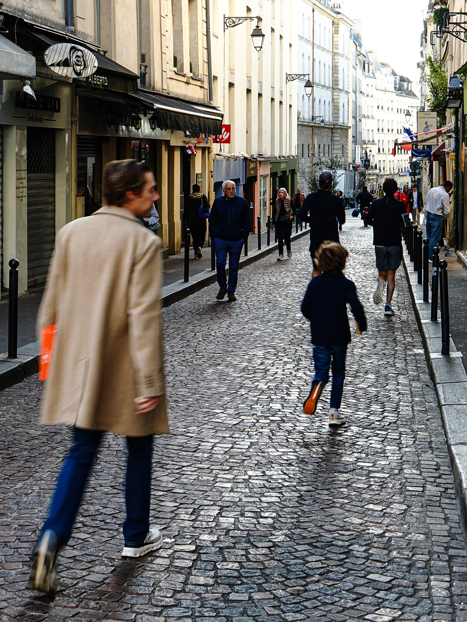 A bustling cobblestone street is filled with people walking between European-style buildings.