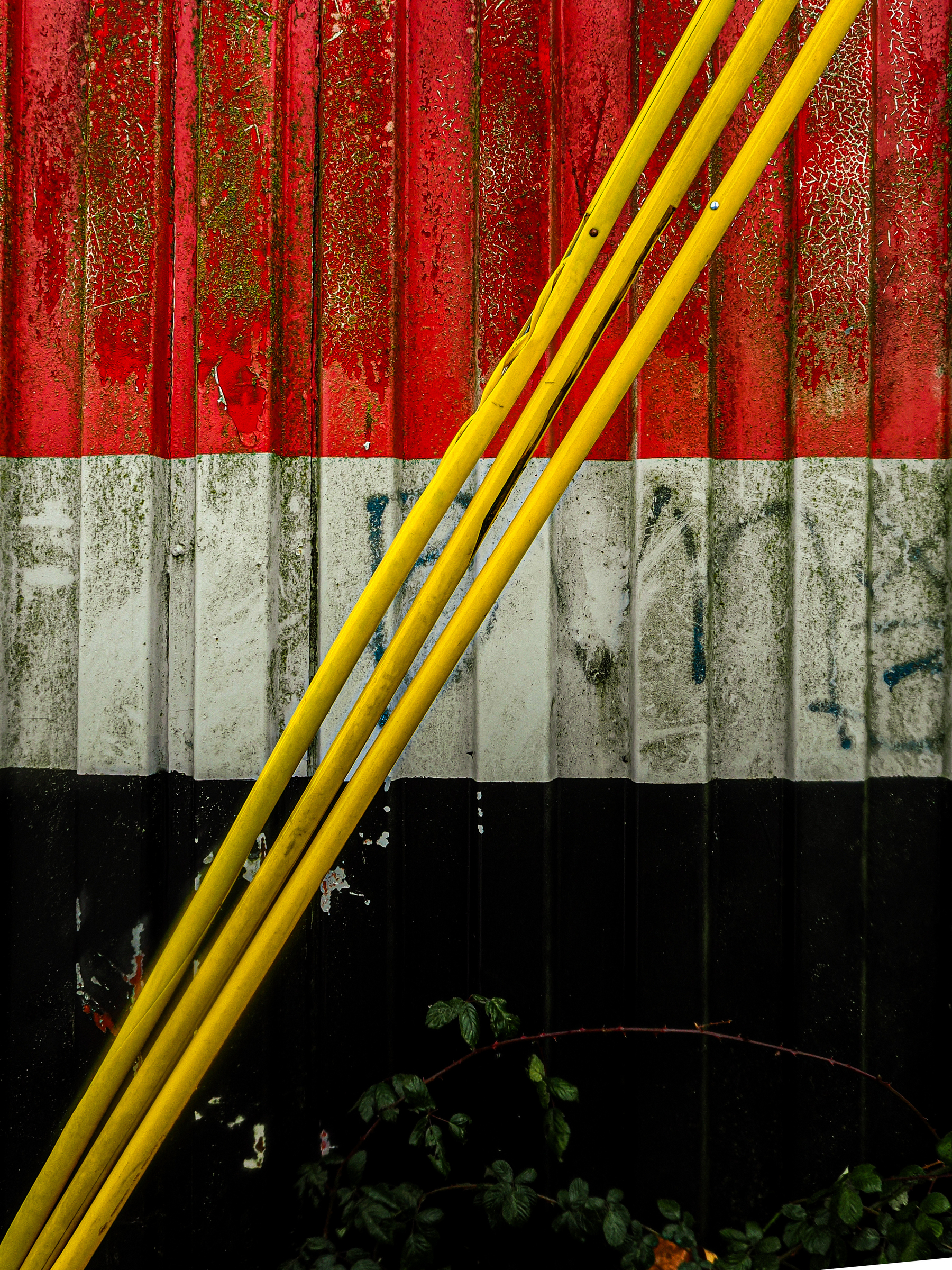 Yellow poles are arranged diagonally across a weathered wall painted with red, white, and black horizontal stripes.