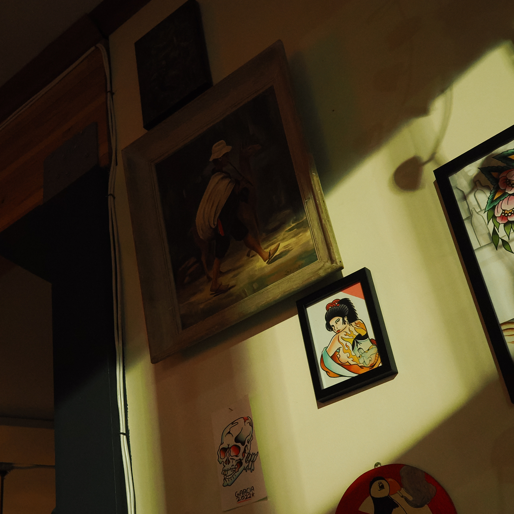 A collection of paintings and artwork, including a portrait of a man walking, a cartoon-like piece, and a skull drawing, are displayed on a shadowed wall.