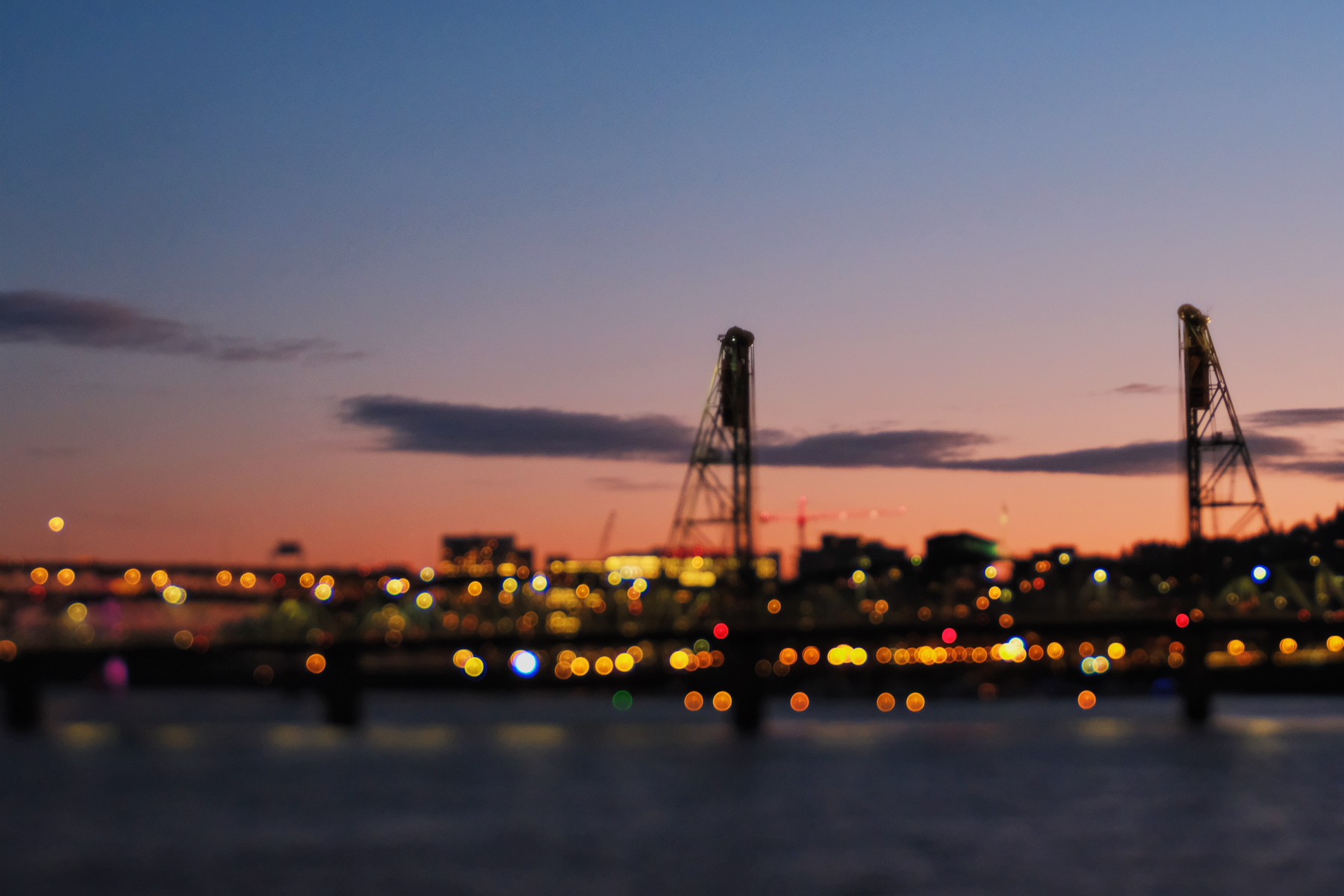 A cityscape at dusk featuring a bridge with illuminated bokeh lights in the foreground and a colorful sky in the background.