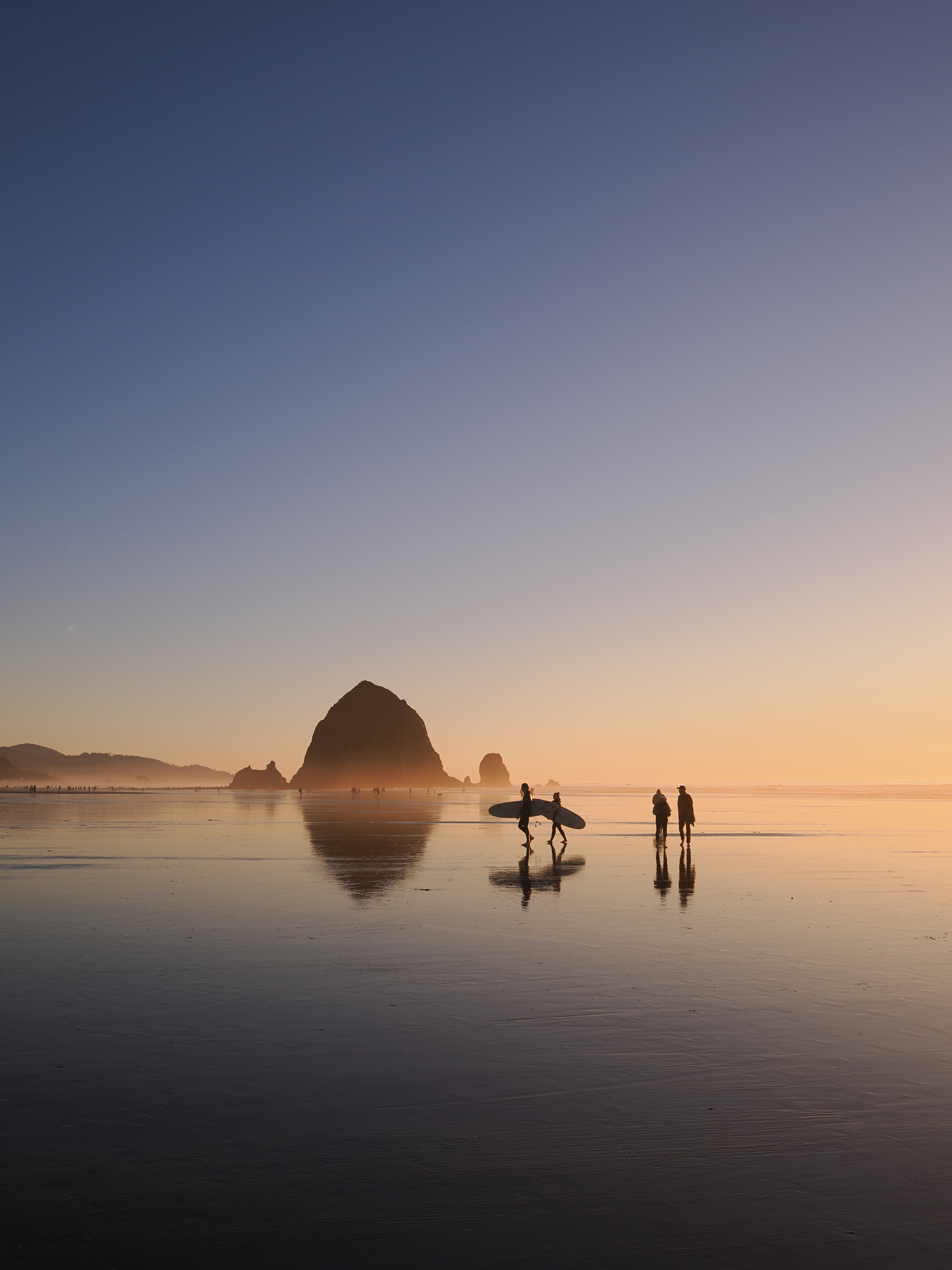 Surfers walk along a reflective, serene beach at sunset with a large rock formation in the distance.